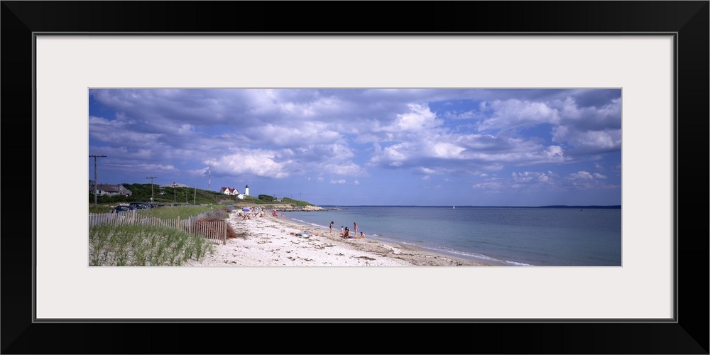 A wide angle photograph is taken of a beach in Cape Cod that only has a few people on it. A light house can be seen in the...