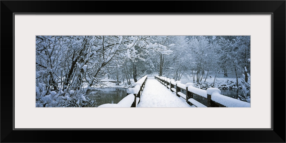 Large panoramic piece of a small bridge that is covered with inches of snow and surrounded by snow covered trees.