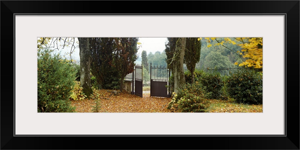 An iron wrought gate is partially open in this rustic garden filled with autumn leaves in a panoramic photograph.