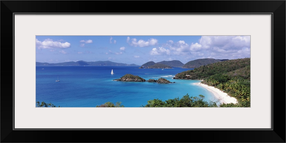 Panoramic photograph of tree filled shoreline with mountains in the distance under a cloudy sky.  There are a few small sa...