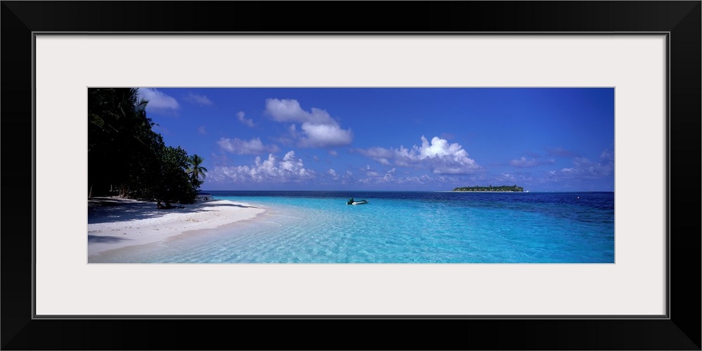 This wide angle photograph is taken of clear ocean water that has a single boat sitting in it with the beach and trees to ...