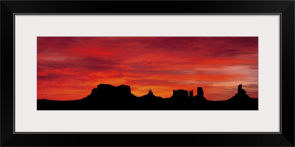 Panoramic photograph of silhouettes of huge rock formations in the desert at sunset under a cloudy sky.