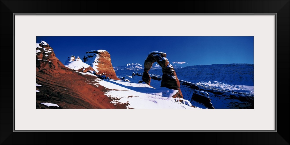 Snow covers desert land and stone arches that are photographed in panoramic view.