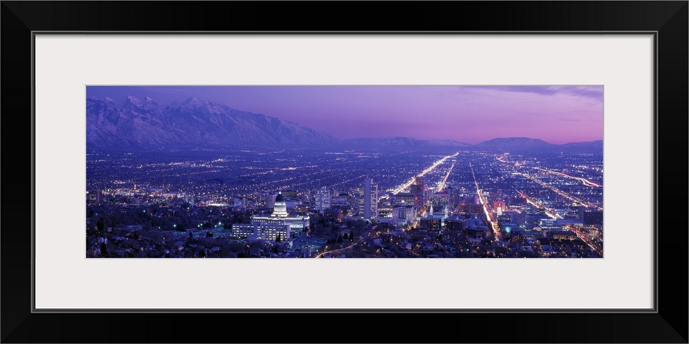 High-angle panoramic photograph of city lit up at dusk with snow covered mountains in the distance under a cloudy sky.