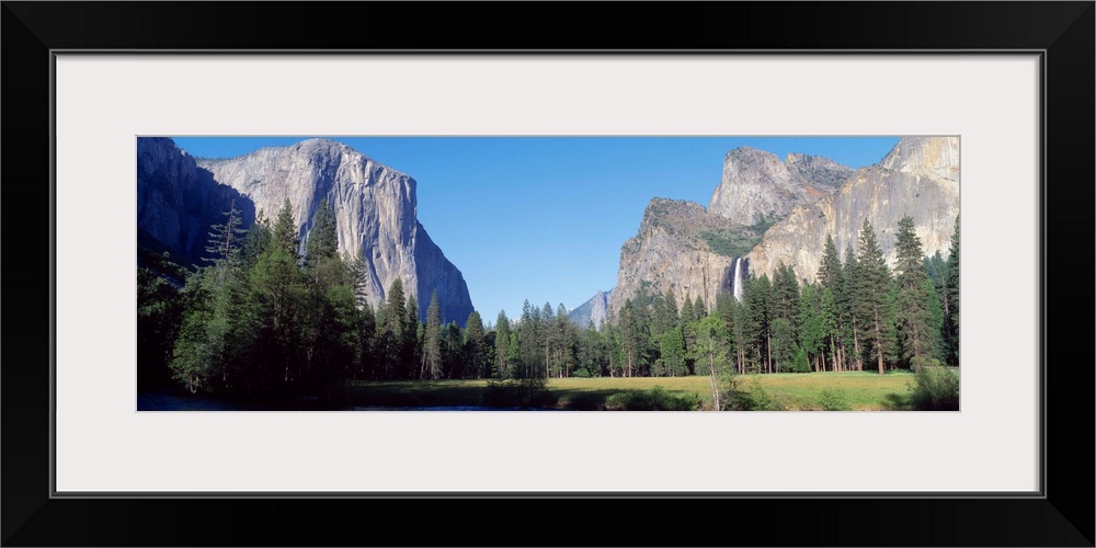 Panoramic photo of large rock cliffs and mountings surrounded by pine trees and water in Yosemite National Park.