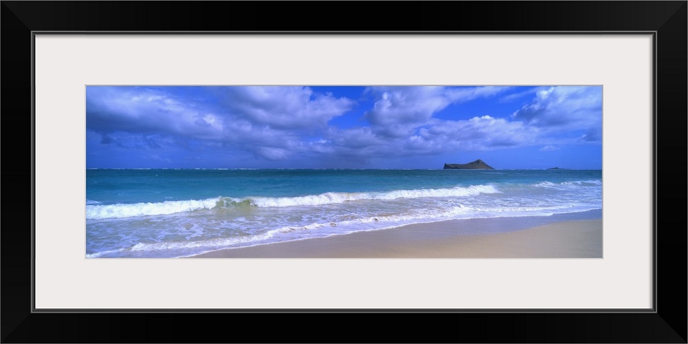 Panoramic photograph of waves breaking on the beach during a cloudy day with island in the distance .