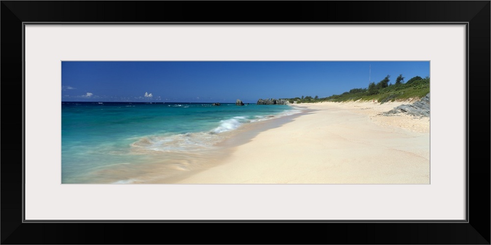 Ocean waves wash up on a sandy tropical shore in this panoramic seascape photographed on a sunny day.