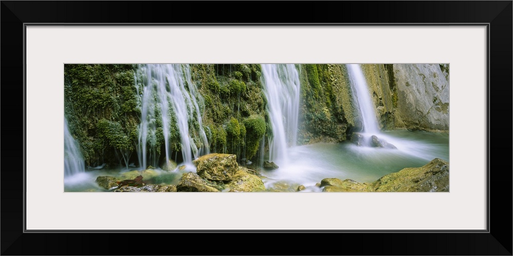 Panoramic photograph taken of several waterfalls cascading over foliage and crashing onto rocks.