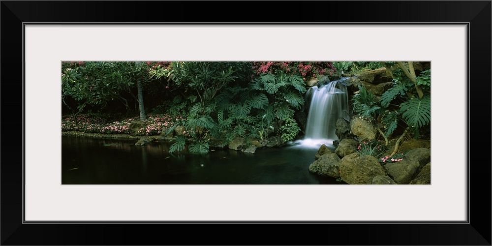 A panoramic photograph of a tropical garden, small cascade tumbles into a circular pool lined with palm fronts and small t...