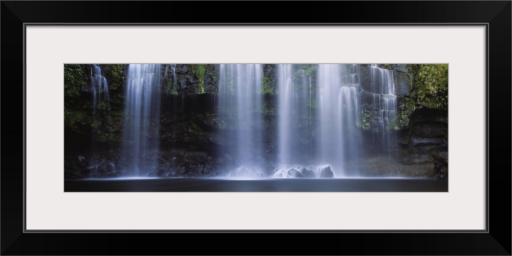 Multiple cascades fall over a moss covered rock face into a pool in this panoramic photograph of a tropical landscape.