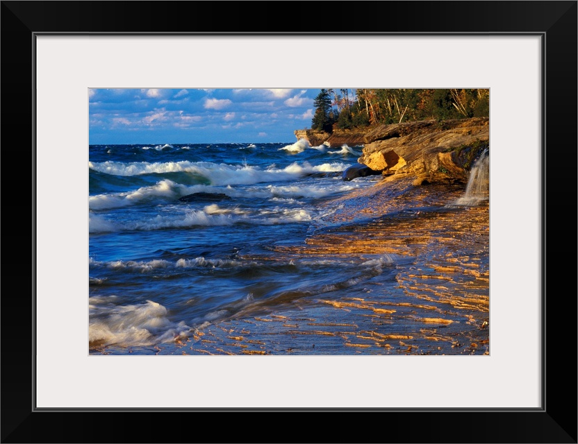 Waves crash onto the rugged lake shore highlighted from the sunset with layered clouds in the distance in this panoramic i...