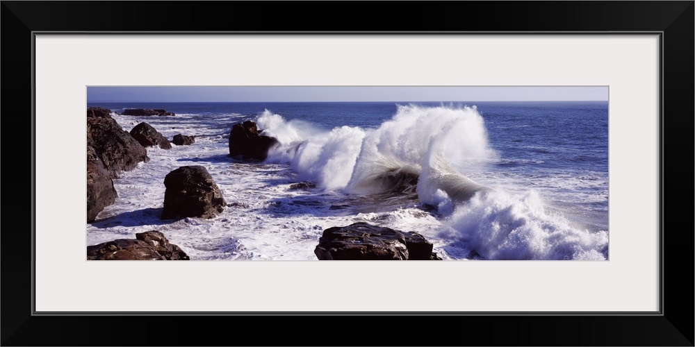 Panoramic photograph of rock cliff line with large boulders and crashing waves under a clear sky.