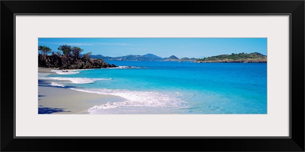 Panoramic photograph displays the calm waters of this bay slowly crashing into the sandy beach, while the mountains in the...