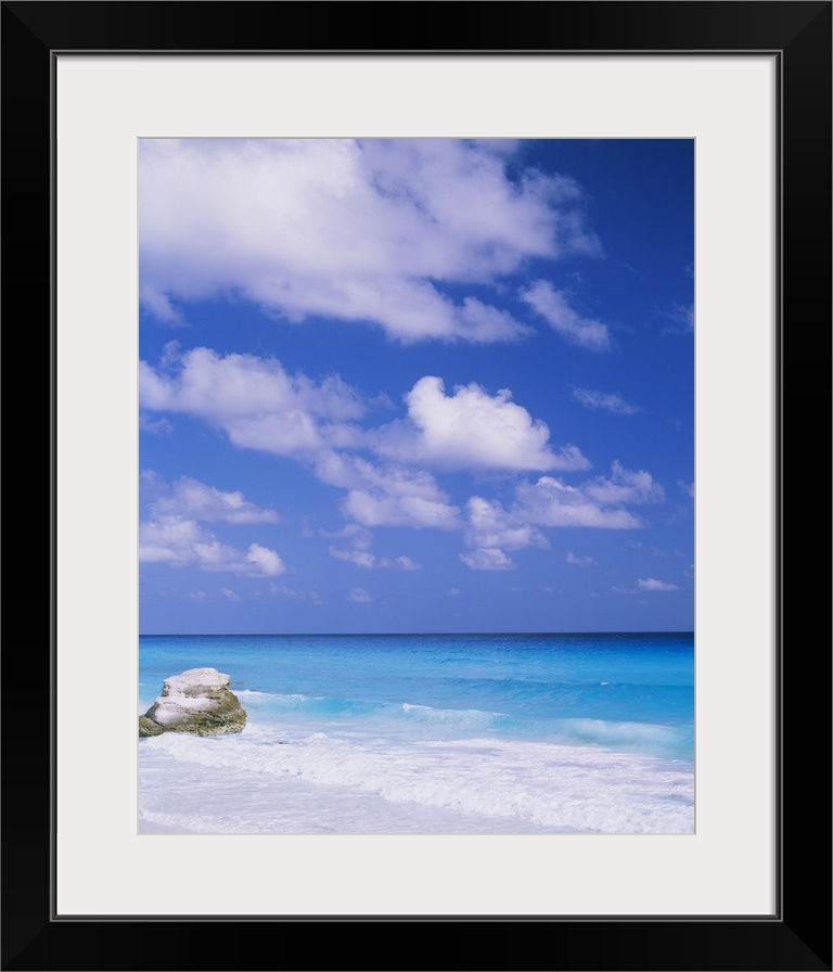 Large photograph displays a lone rock sitting on a beach, while the waves of a sea begin to crash against it and the sandy...