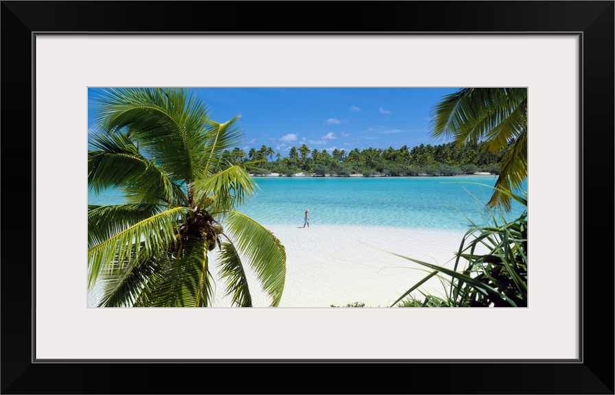 Photograph of a lady walking alone on a white sand beach surrounded by crystal clear water and palm trees.