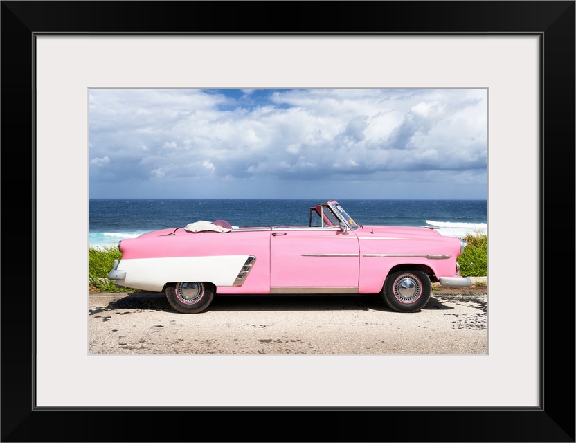 Photograph of a light pink and white vintage convertible parked in front of the ocean.