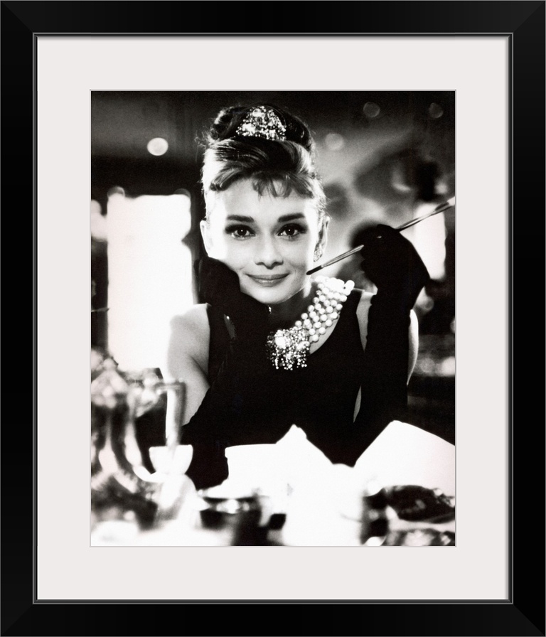 Classic iconic photograph of Audrey Hepburn sitting at a table holding a cigarettte and smiling.