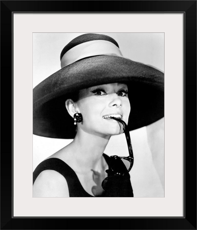 Large photograph of Audrey Hepburn in a giant sun hat holding sunglasses in her mouth as she glances at the camera in a bl...