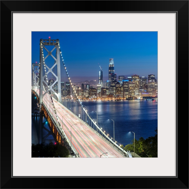 Square photograph of the Bay Bridge at dusk with downtown San Francisco lit up in the background.