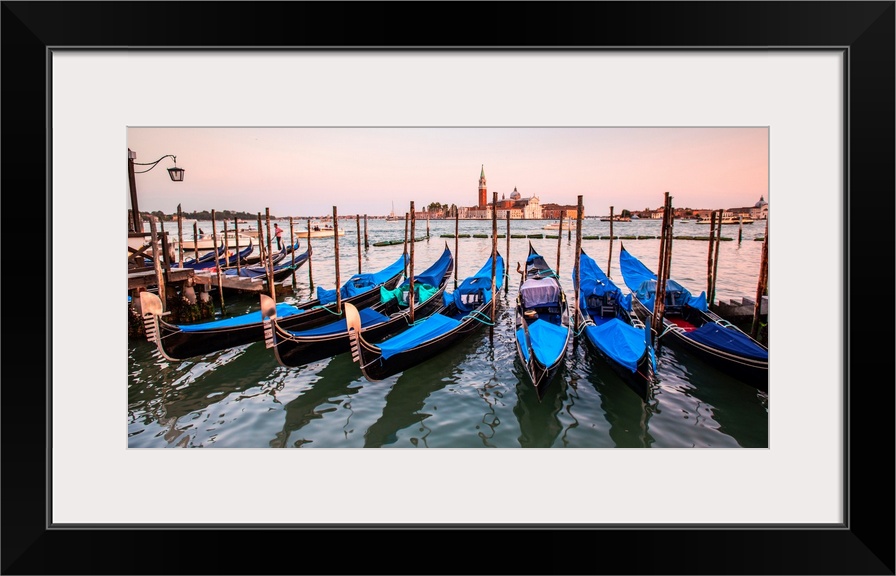 Panoramic photograph of blue gondolas docked in a row on the water with St. Mark's Square in the background at sunset.