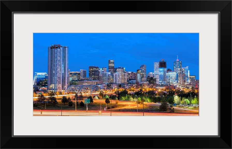 Photograph of the Denver, CO skyline at dusk with warm light trails on the highway from passing cars.