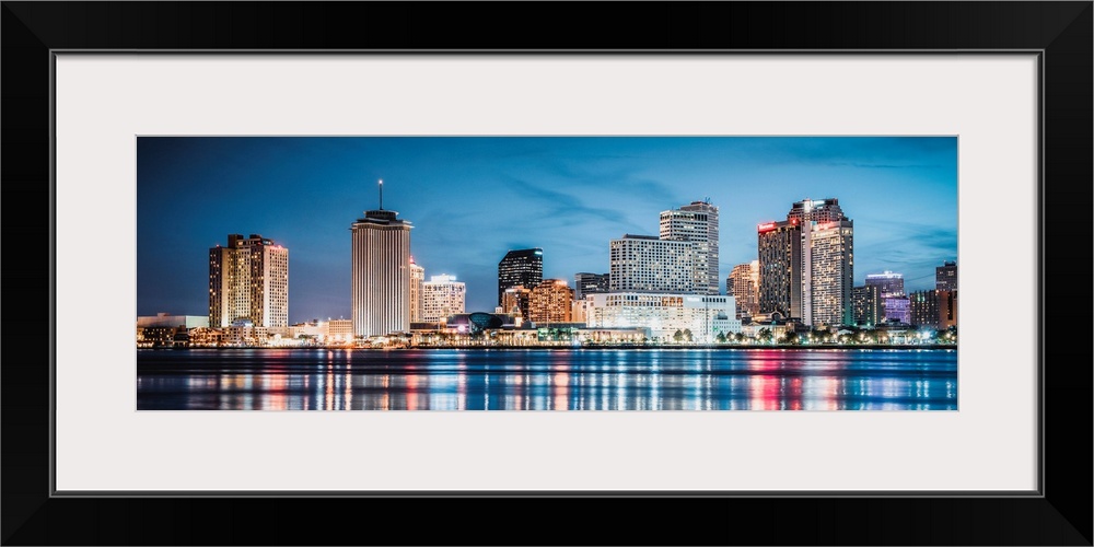 Panoramic photograph of the New Orleans skyline lit up at dusk and reflecting colorful bands onto the Mississippi River.