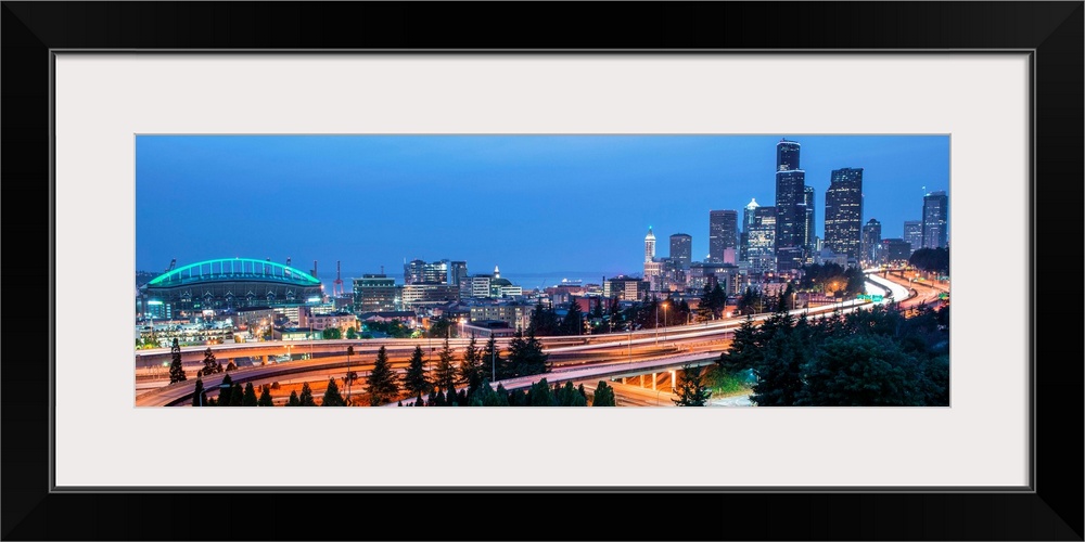 Panoramic photograph of the Seattle skyline with the stadium on the left and light trails from traffic on the highway.