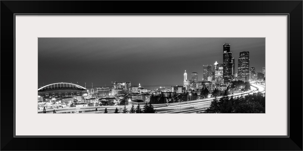 Panoramic photograph of the Seattle skyline with the stadium on the left and light trails from traffic on the highway.