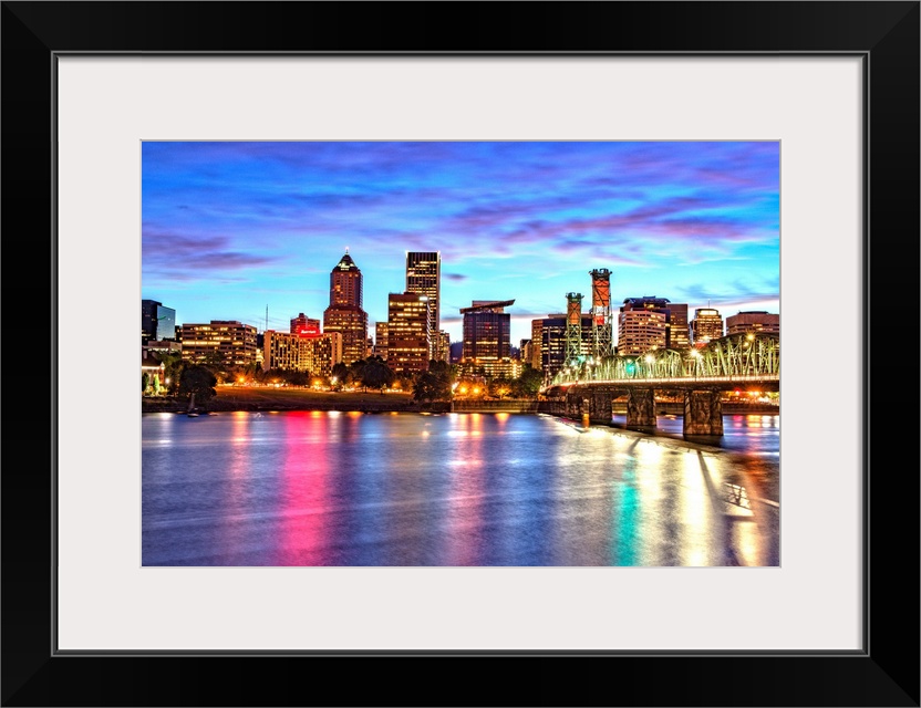 Photograph of the Portland, Oregon skyline lit up at sunset with colors reflecting onto the Willamette River in the foregr...