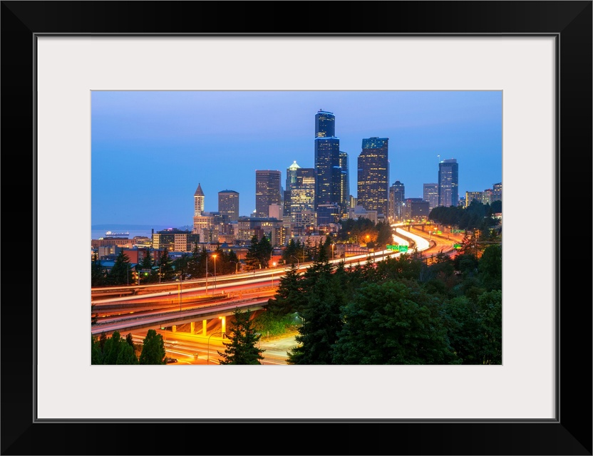 Photograph of the Seattle skyline at dusk with light trails from the cars on the highway in the foreground.