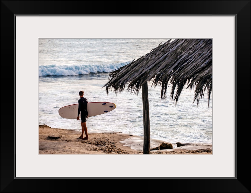 Photograph of a surfer walking along the shore of the pacific ocean in San Diego, California, with a cabana built with nat...