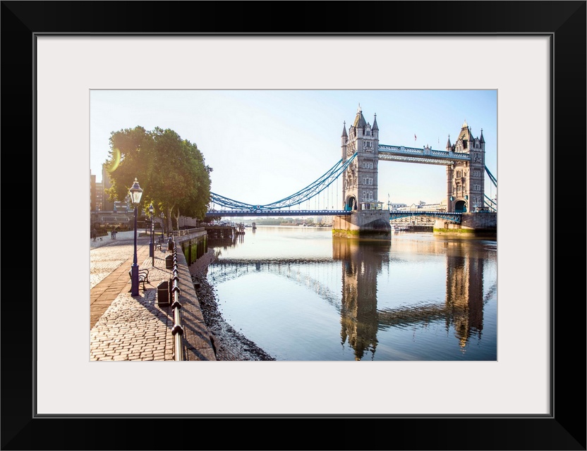 Photograph of Tower Bridge  reflecting on River Thames with a brick sidewalk on the side.