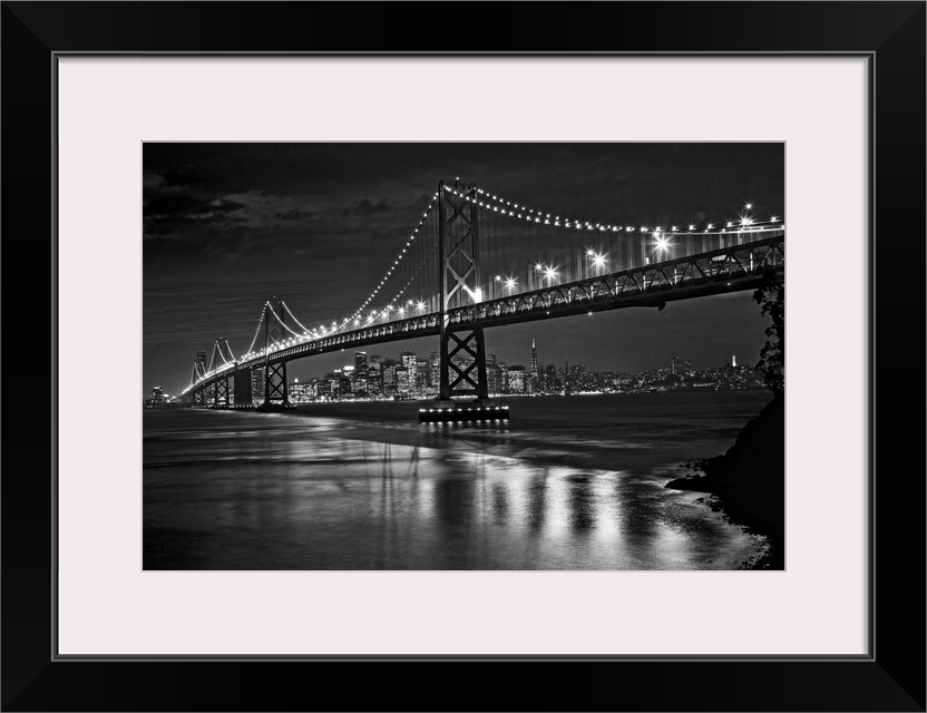 This landscape is a high contrast, monochromatic photograph that shows a bridge in the fore ground and glowing city skylin...