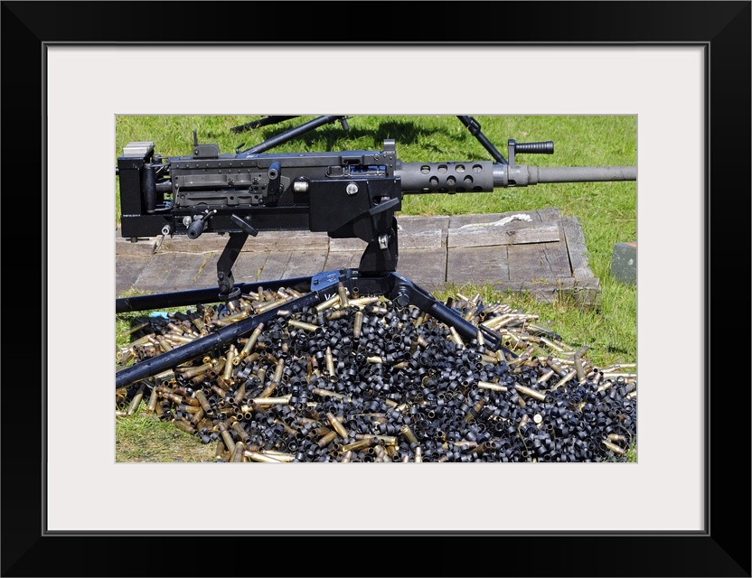 Big, horizontal, close up photograph of a .50 caliber browning machine gun, sitting in the grass, over a large pile of use...
