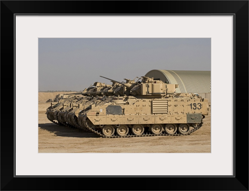 Large landscape photograph of a line of M2/M3 Bradley Fighting Vehicles cruising over a dirt landscape, a single building ...