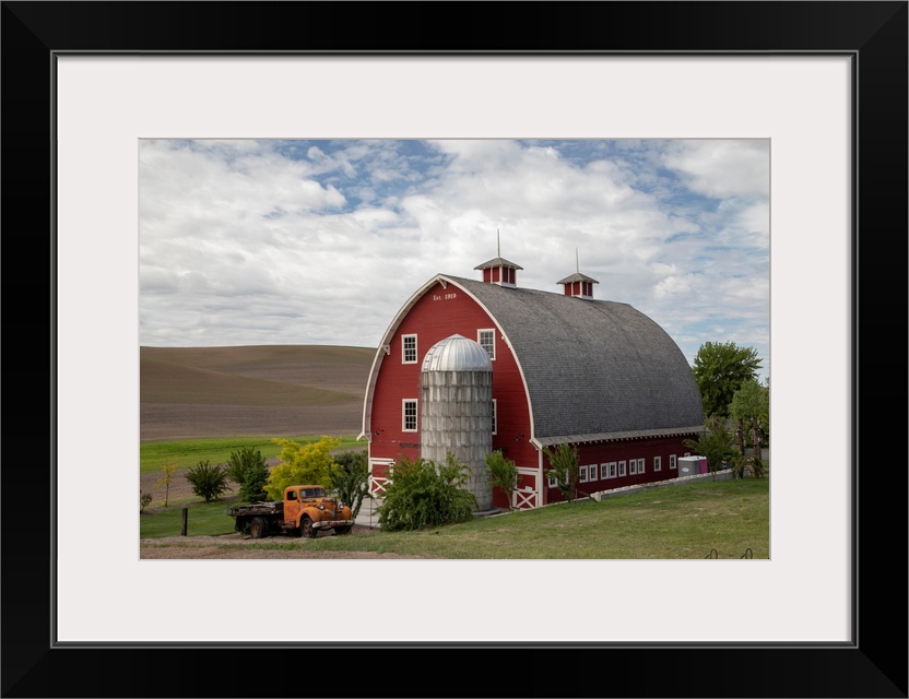 A photograph of a large red barn and grain silo with a vintage orange truck parked out front