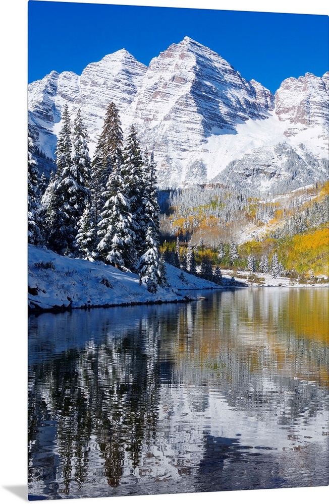 Snow covered mountains are pictured against a clear blue background that reflects down in the body of water that is in the...