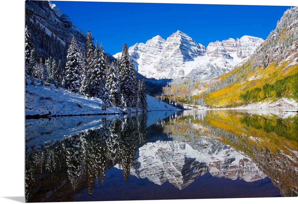 A snow covered mountain range with tall trees in front reflects beautifully into a body of water.