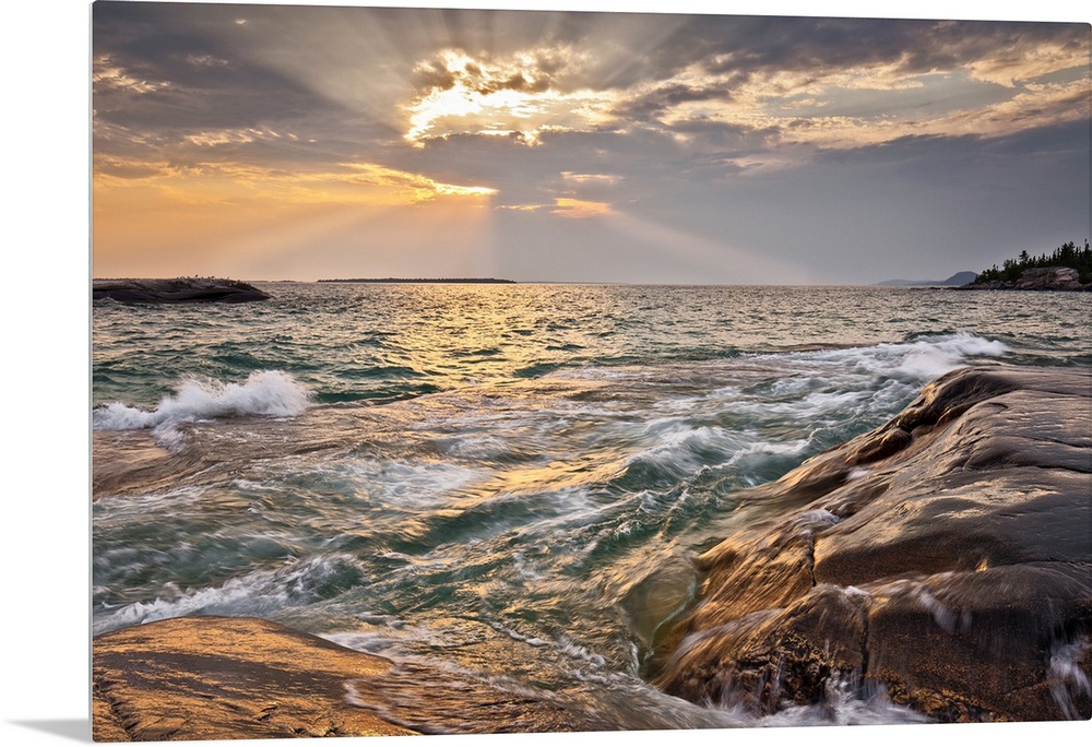 Burst of sun rays peeking out behind a cloud as waves lap on to a rocky shore.