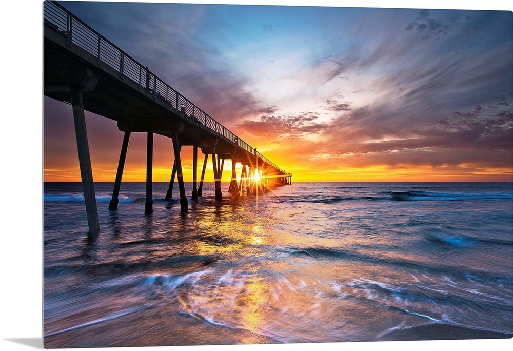 Sun intersects pier at sunset with colorful waves and sky.