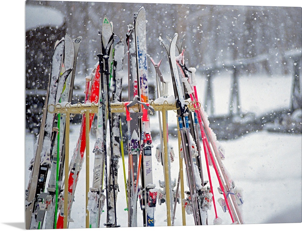 In this photograph multiple skis are stored on an outside rack as large white snowflakes fall all around. Blurred backgrou...
