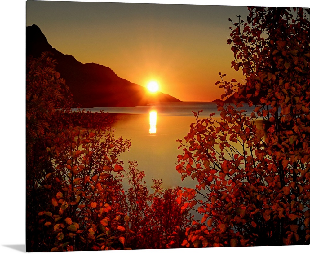 Big photograph emphasizes the sun as it begins to set over a mountain and then reflect over a calm body of water in Norway...