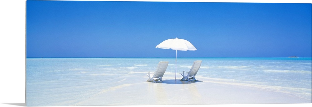 A panoramic photograph displaying two chairs sitting underneath an umbrella on a sandy beach in Maldives.  The clear water...
