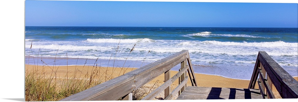 Panoramic photo of a wooden walkway leading to a beach shore with crashing waves during the day.