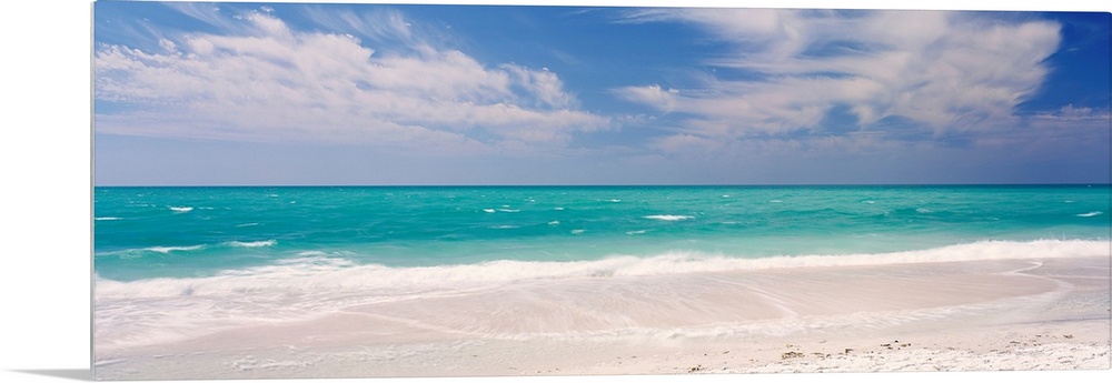 Panoramic photograph of calm ocean with surf and sand in the foreground and cloudy sky above.