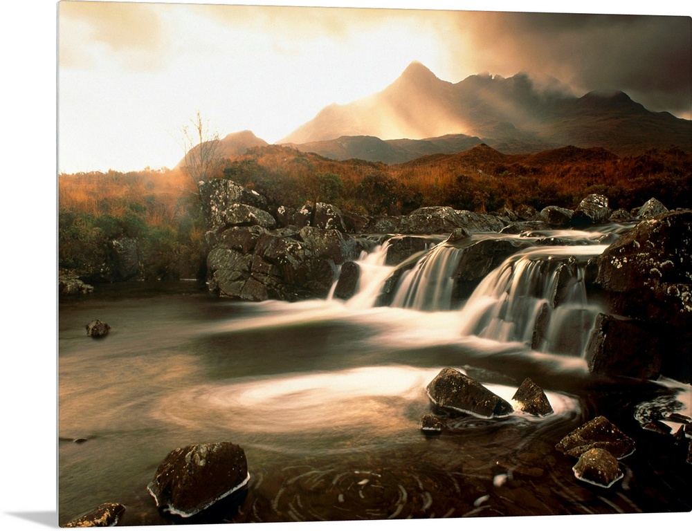 Time lapsed photograph of water flowing through a rocky river while the sun glows behind clouds in the distanced.