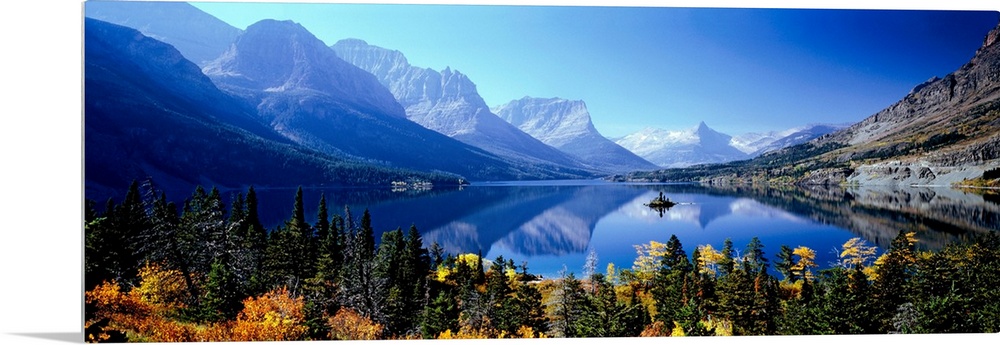 A panoramic photograph of mountains reflecting in a still lake surrounded by trees in this landscape of the wilderness.