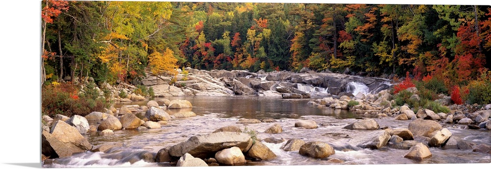 A relaxed panoramic landscape of large boulders in a New England river lined with trees with autumn colors.