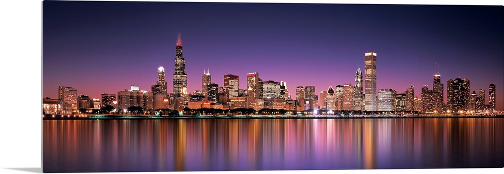 Panoramic photograph of the Chicago, Illinois skyline filled with skyscrapers reflecting onto Lake Michigan at night.
