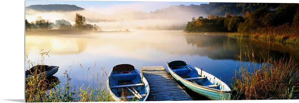 Panoramic photograph showcases rowboats sitting next to a dock on a lake in Grasmere within Cumbria, England.  In the back...